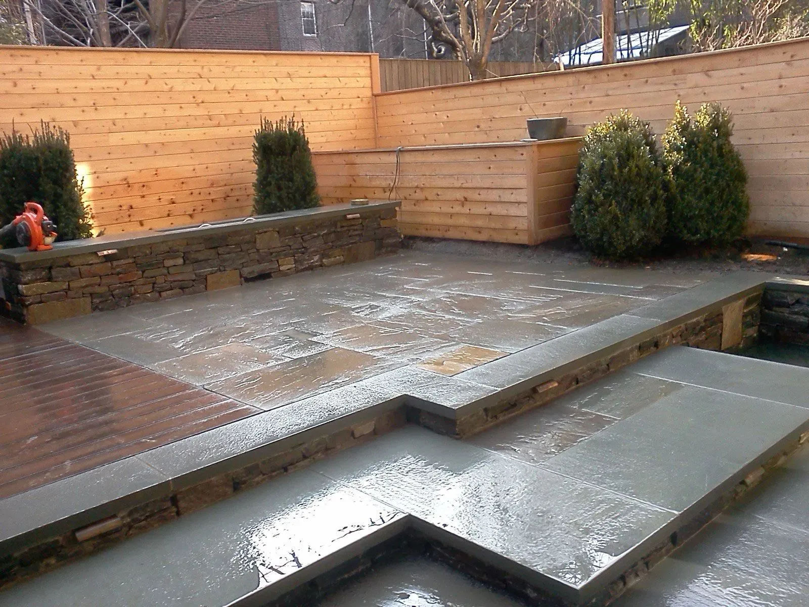 Backyard patio with wet concrete steps, wooden decking, stone wall, and shrubs after rain