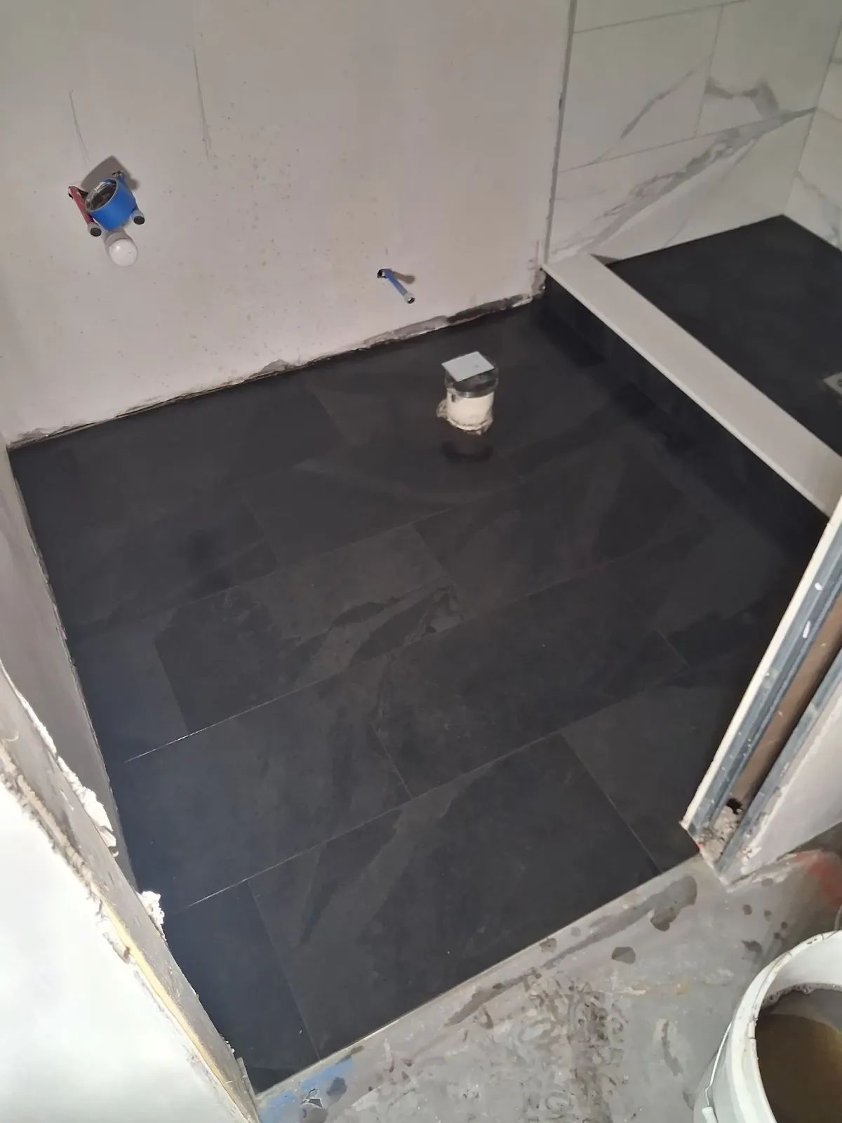 Small tiled room under renovation with black floor tiles, exposed plumbing, and a step in the corner.