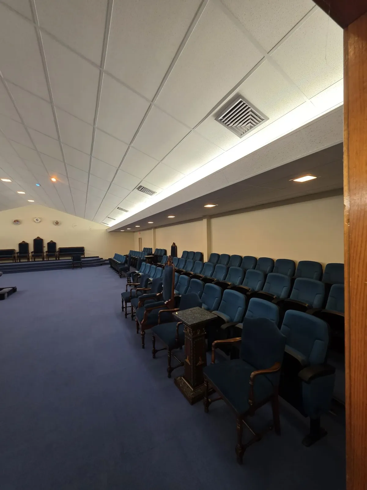 Empty auditorium with rows of blue seats and a carpeted aisle under a slanted ceiling.