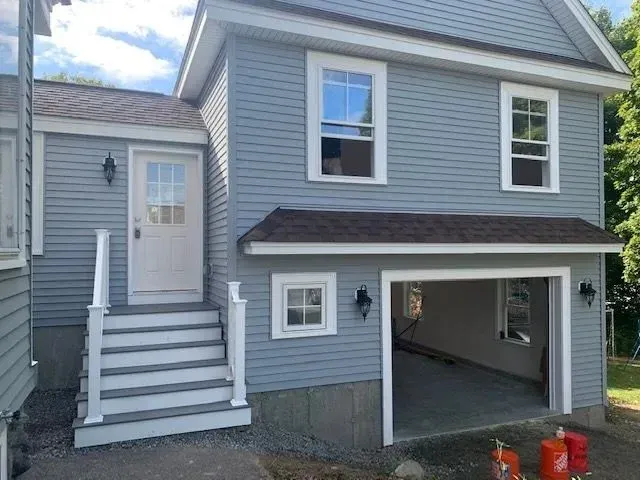 Front of a two-story house with blue siding, white trim, and a garage with an open door. There are stairs leading up to a side door with a small porch area, and several windows on both stories. Outdoor lighting fixtures are visible near the entrance