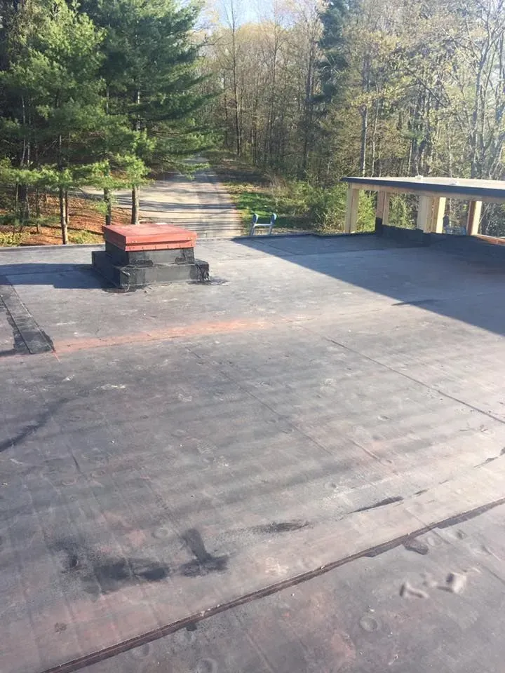 Flat black roof with red chimney and wooden deck in a wooded area.