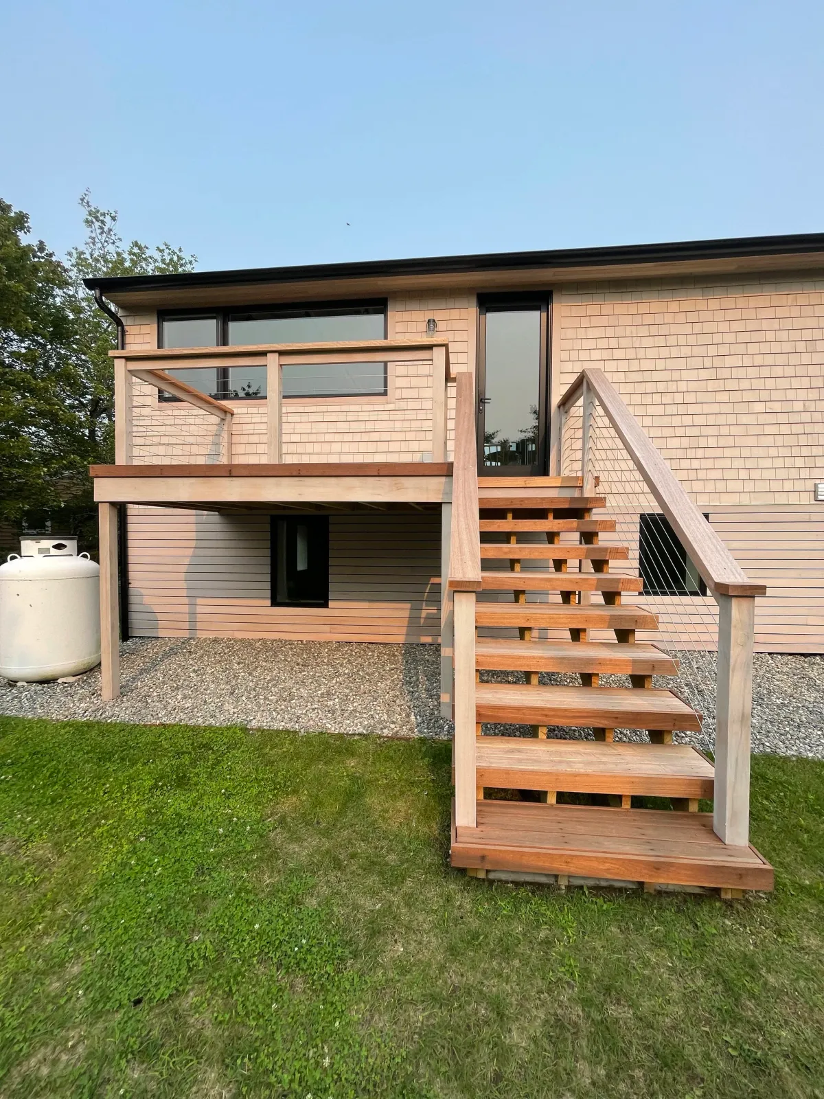 Backyard view of a two-story house featuring a wooden deck and staircase, with a grassy lawn and a propane tank on the left.