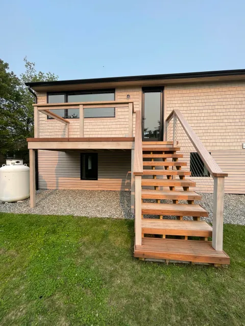 Backyard view of a two-story house featuring a wooden deck and staircase, with a grassy lawn and a propane tank on the left.