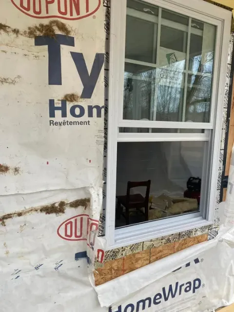 A new white window installed in a wall covered with Tyvek HomeWrap house wrap, showing exposed wooden framing at the sill.