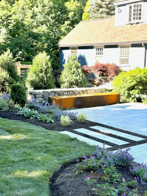 A stone patio and attendant landscape design create an exterior haven with trough style fountain and adjacent gardens.