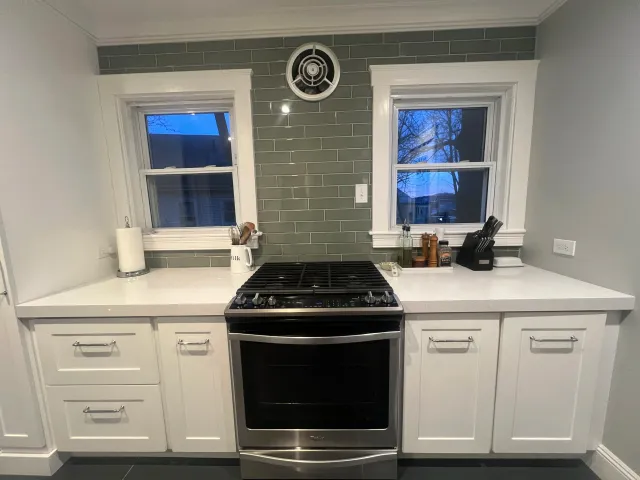 Kitchen with white cabinets, a stove, gray tiled backsplash, and two windows with trees outside at dusk. Installed by Total Home Services