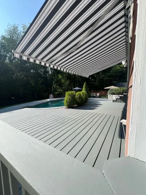 View of a backyard with a swimming pool, wooden deck, and outdoor furniture, partly shaded by a striped retractable awning.