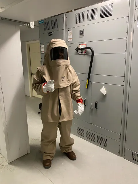 Person wearing a beige hazmat suit, gloves, and face shield standing in front of electrical equipment.