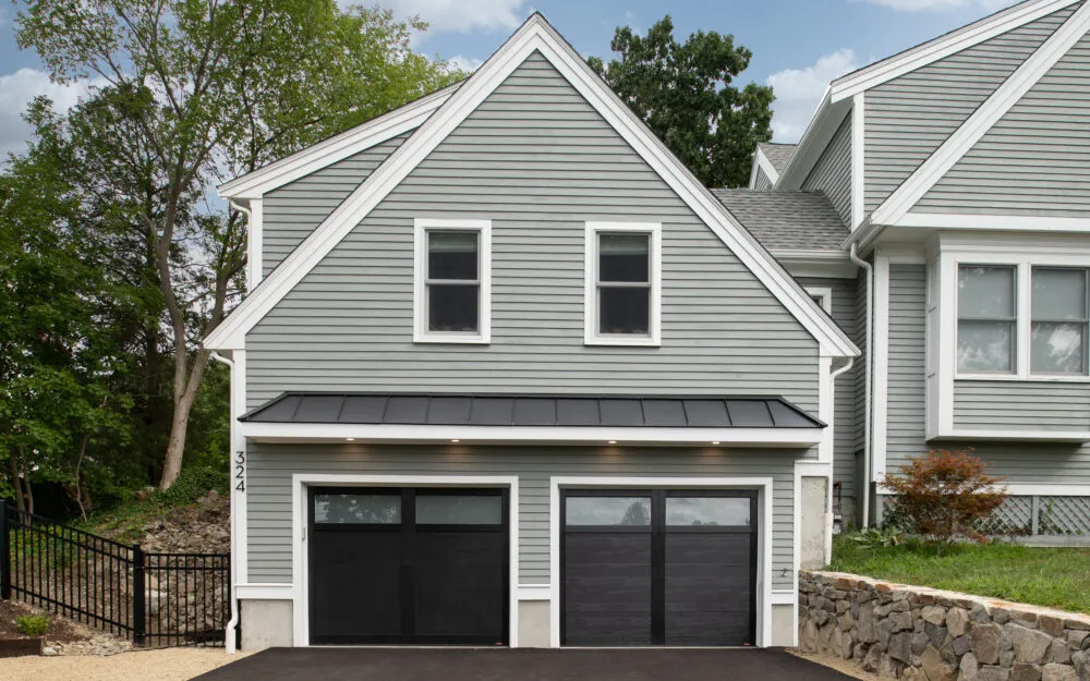Modern gray remodel with white trim, black garage doors, trees, and stone landscaping – Woodland Builders, South Shore MA.
