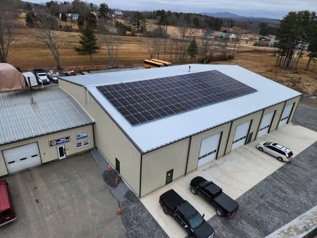 Large metal warehouse with solar panels on the roof, multiple garage doors, parked vehicles, rural setting.