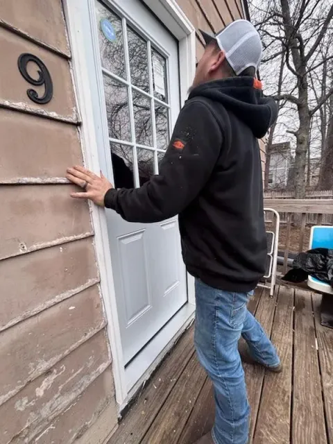 Man in jeans and a hooded sweatshirt leans against a white door with a window, on a wooden deck.