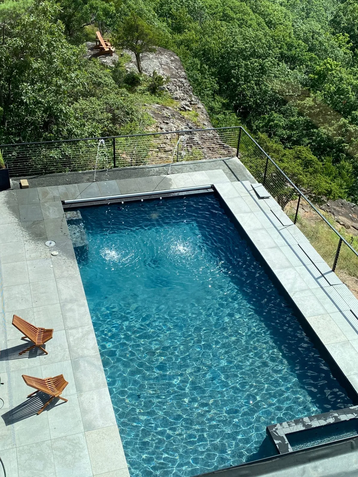 Modern residential pool with tile deck, water features, and wooded backdrop