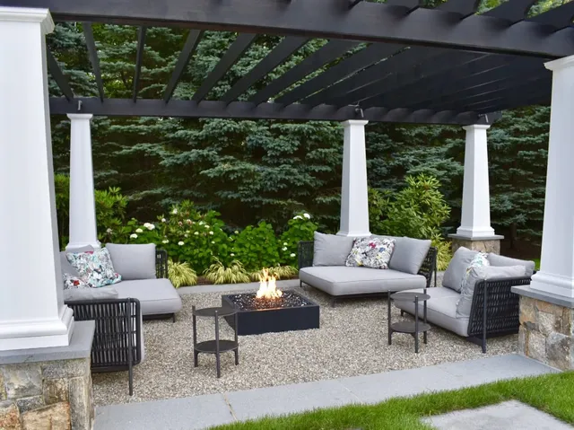 An outdoor patio area with a pergola, comfortable seating, decorative cushions, a fire pit, surrounded by greenery and landscaped with pebbles.