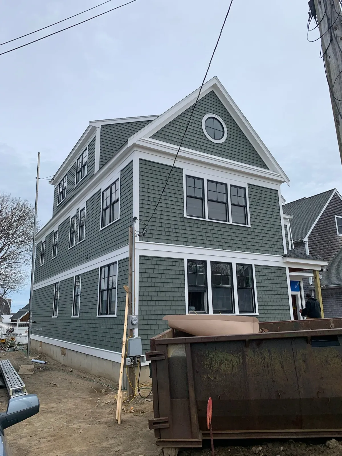 Three-story house with green siding and white trim, several large windows, and a pitched roof with decorative round window, under construction, with a worker near the entrance, a large dumpster in the foreground, and power lines overhead. Scituate.