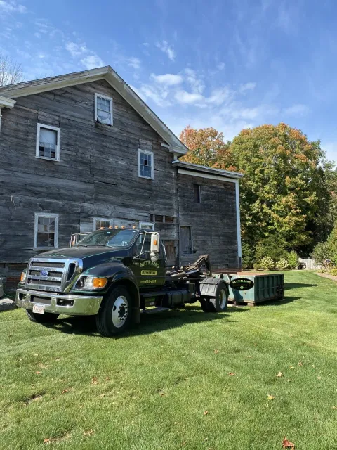 Black dump truck next to weathered wooden house, green lawn, sunny day.