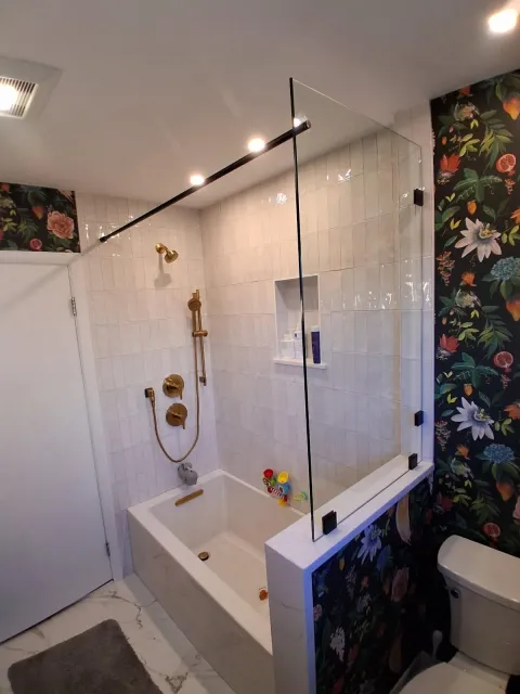 Bathroom with glass shower screen, white tub, gold fixtures, white tile, and floral wallpaper.
