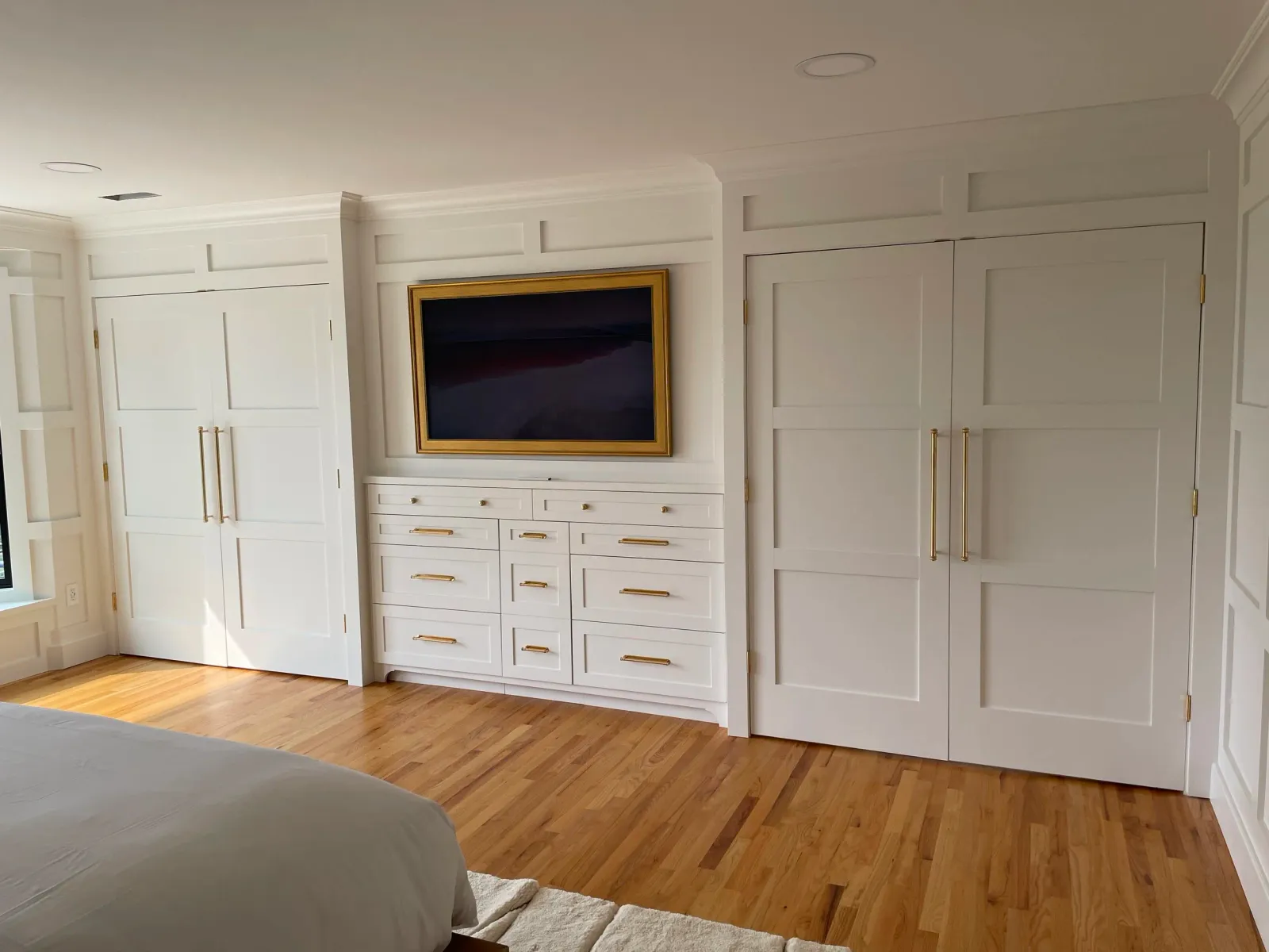Bedroom with white built-in cabinets, a wall-mounted television with a gold frame, and hardwood floors.