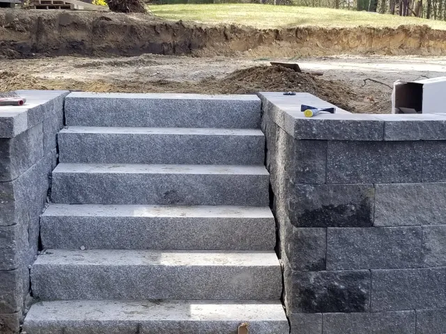 Gray stone staircase with four steps leading up to a level ground, flanked by stone walls, at a construction site with exposed dirt and trees in the background.
