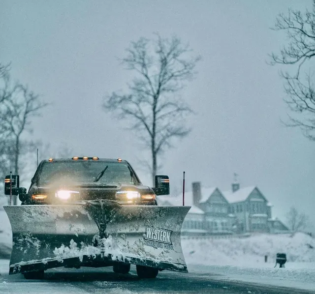 Truck with a Western snow plow clearing snow from a residential driveway in winter, provided by Perfect North Lawn Care in New Hampshire.