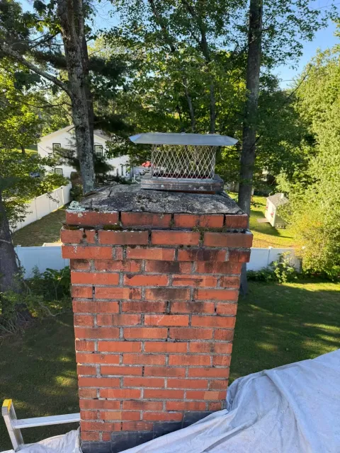 A brick chimney with a metal cap atop a house roof, surrounded by green trees and a white fence in the background.