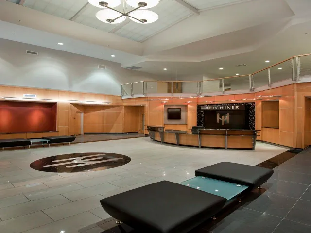 Empty hotel lobby with a reception desk, seating area, flat-screen TV, wood paneling, and a large decorative logo on the floor.