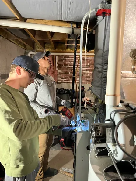 Two workers using a welding torch in a basement utility area with exposed pipes and ductwork