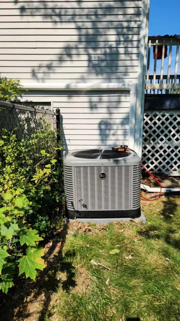 Outdoor air conditioning unit next to a house wall, with a fence on the side and some bushes in the foreground.