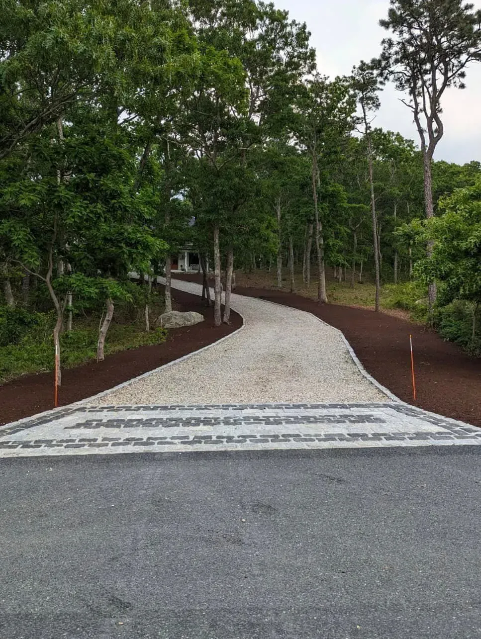 Gravel driveway with patterned border leading through a wooded area.