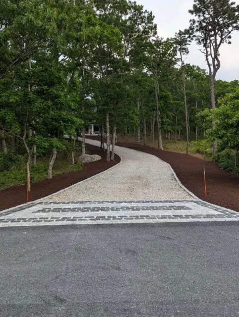 Gravel driveway with patterned border leading through a wooded area.