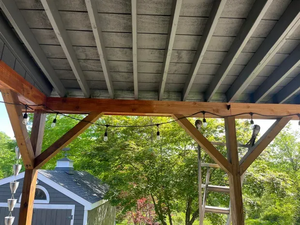 Wooden pergola with string lights, attached to a gray-painted ceiling. Green trees in the background.