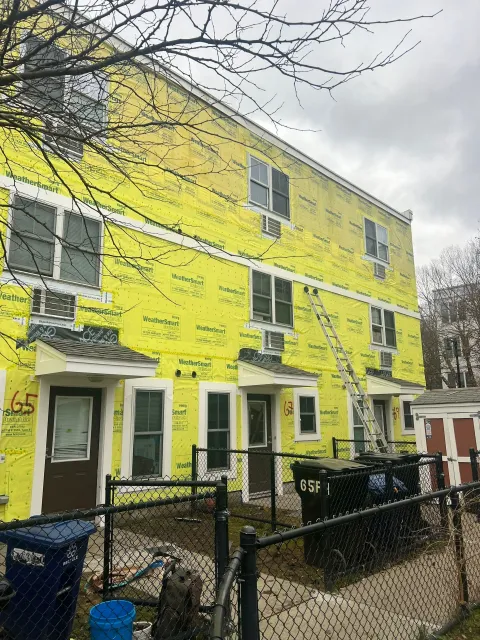 The Franklin Hills Apartments under construction. A yellow-wrapped building, windows, doors, ladder, trash bins, and fence in overcast conditions.