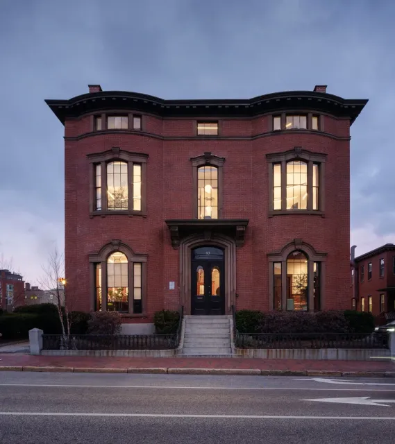 A large, historic red-brick building designed by a Maine architect features tall, lit windows and arched doorways on a quiet city street at dusk.