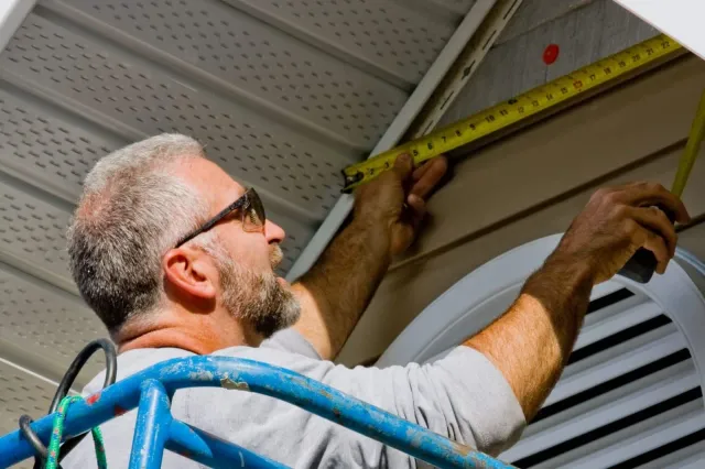 A man with gray hair and a beard, wearing glasses, measuring a section of a ceiling with a yellow tape measure.