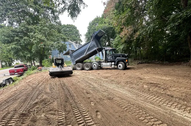A black dump truck in a dirt clearing, with its bed raised to dump soil next to trees under a bright sky.