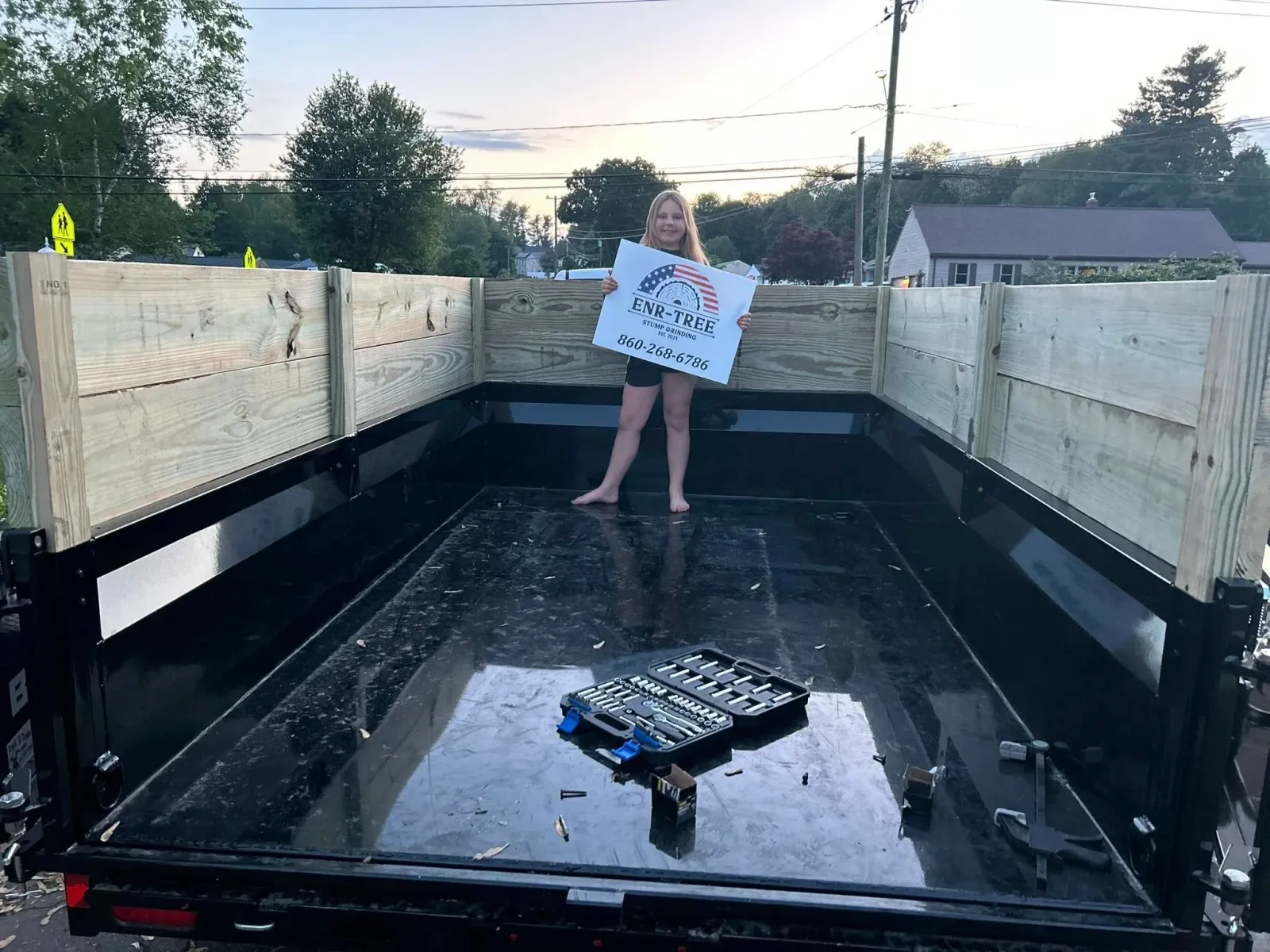 A young girl with long blonde hair, barefoot, holding a sign that reads 'ENR-Tree Stump Grinding' with a phone number, stands inside a trailer with tall wooden side panels. The trailer is parked outdoors in a suburban neighborhood during early evening, with houses, trees, and utility poles visible in the background. There are tools and equipment on the trailer floor.