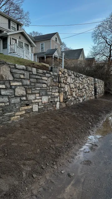 Stone retaining wall beside muddy path, with houses on a hill under a blue sky.