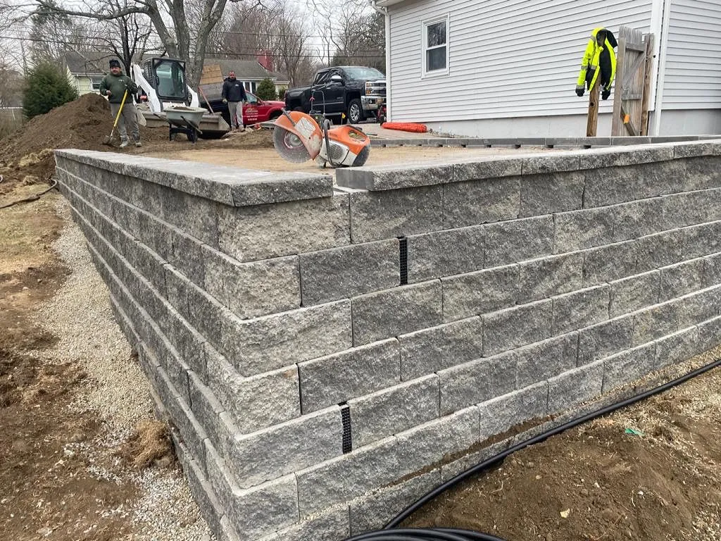 Retaining wall construction using gray interlocking blocks; tools and workers are visible in the background.