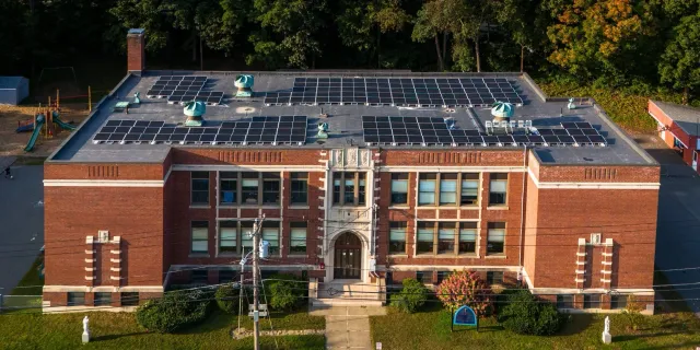 Aerial view of a brick school building with a flat roof covered in solar panels, surrounded by trees and playground equipment.