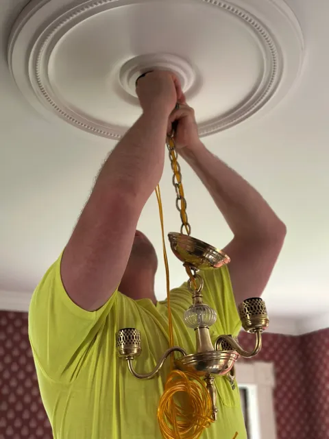 A person changing a ceiling light fixture, with a chain hanging down, in a room with red patterned wallpaper.