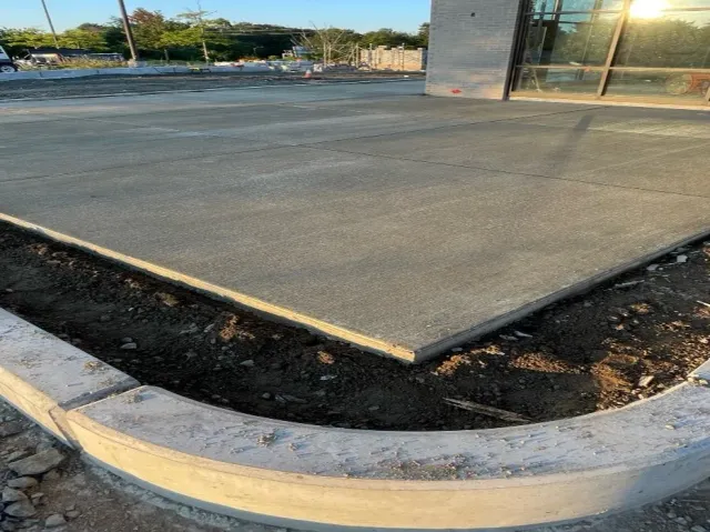 Freshly poured concrete sidewalk at a construction site, with a curved edge and a building with glass windows nearby.