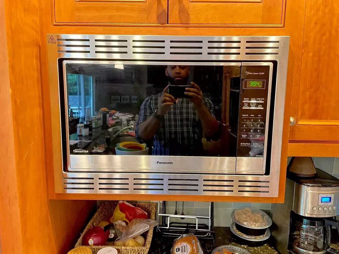 Stainless steel microwave built into wooden kitchen cabinets, reflecting a person taking the photo.