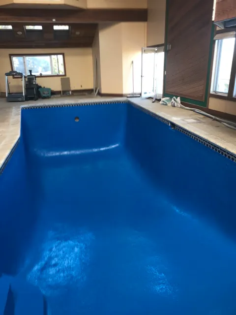 Empty indoor pool with a blue finish, under construction or renovation, with tools and construction materials nearby, in a room with windows and wooden ceiling.