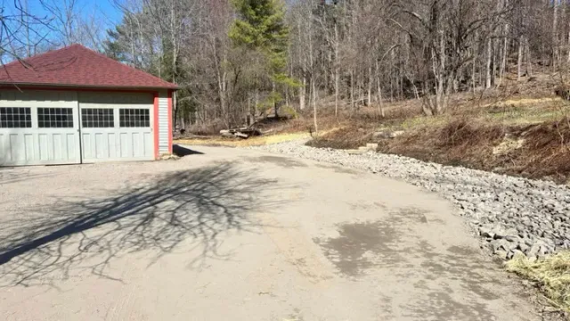 A cream-colored garage with a red roof stands next to a gravel driveway bordered by stones and a forest in the background.