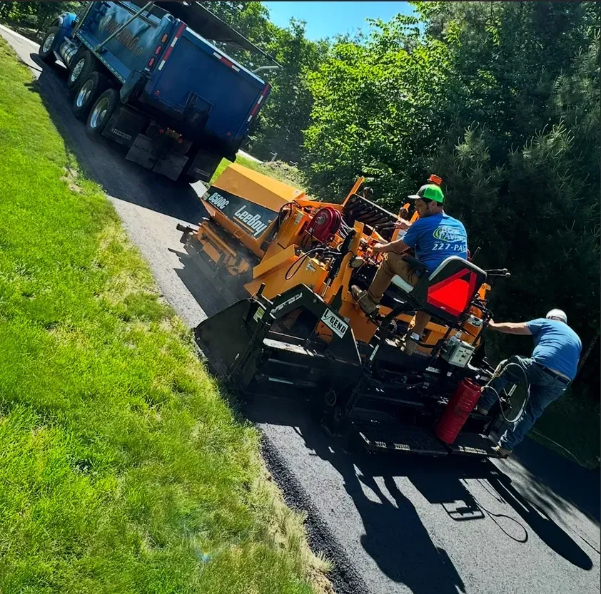 Workers operate heavy machinery to pave a road with asphalt, with a dump truck nearby in a sunny, outdoor setting.