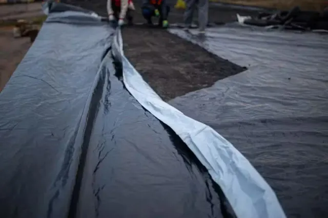 Close-up of a black plastic sheeting or tarp laid on the ground, with a folded silver reflective material along one edge, in an outdoor setting with people and other materials in the blurred background.