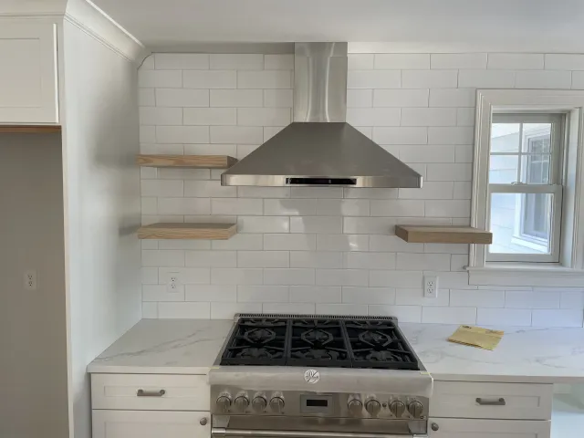 Modern kitchen with white cabinets, marble countertops, stainless steel stove and vent hood, white subway tile backsplash, and two open wooden shelves near a window.