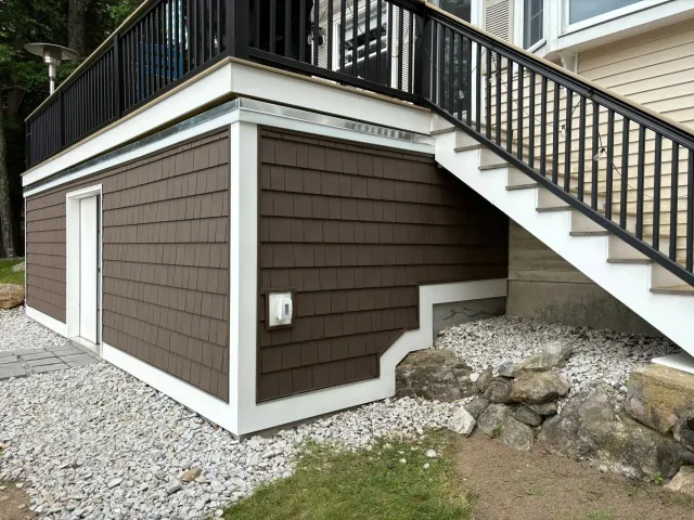 Exterior view of a house with brown siding, a staircase with black railings, and a small enclosed area at the bottom of the stairs, surrounded by rocks and gravel.
