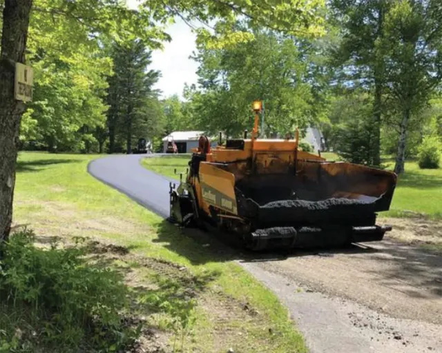 Asphalt paving machine laying fresh asphalt on a driveway, with trees and a house in the background.