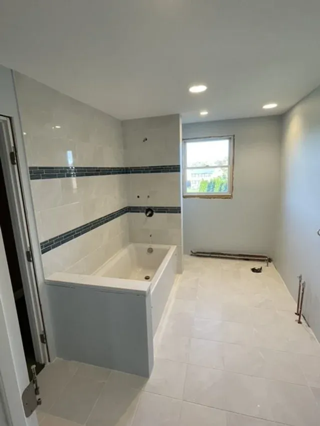 Bathroom under renovation with a partially installed bathtub, tiled walls with horizontal black and gray tiles, a window, and no fixtures installed yet.