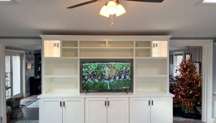 Floor-to-ceiling built-in media unit in living room, housing flat-screen TV flanked by open shelves and closed cabinets, accented with recessed lighting; ceiling fan above, Christmas tree visible in adjacent space.